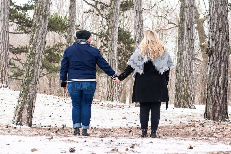 Weight bias affects health, especially when it comes from healthcare providers. Image is of two young women in larger bodies, standing in a forest holding hands, with their backs towards the camera. One woman is wearing jeans, a blue casual jacket, and a black stocking cap. The other woman is wearing black skinny jeans, a black knee-length coat, and a large light gray blanket scarf around her shoulders, under her long blonde hair.