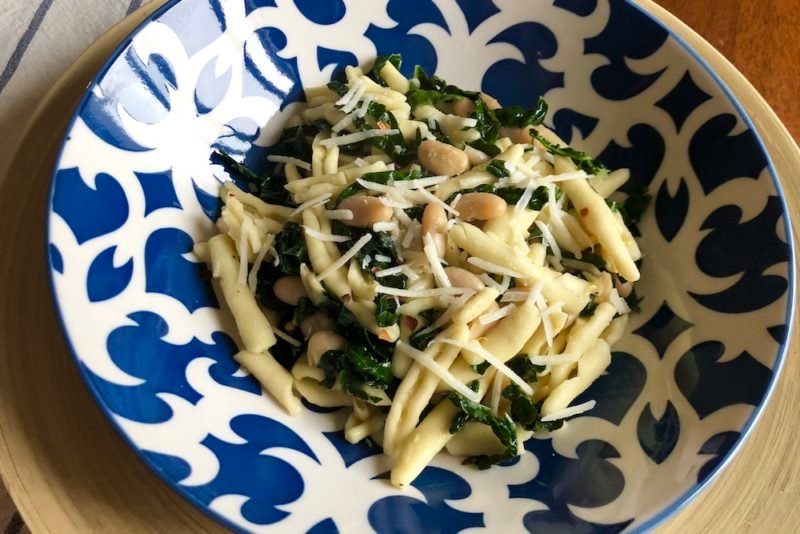 Photo of a pasta dish with kale, white beans and Parmesan cheese, served in a cobalt blue and white pasta bowl on a bamboo charger.