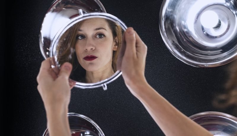 Beauty myth. Photo of a woman holding a large, round makeup mirror at arm's length. We see her face reflected in the mirror. She has long brown hair and red lipstick.