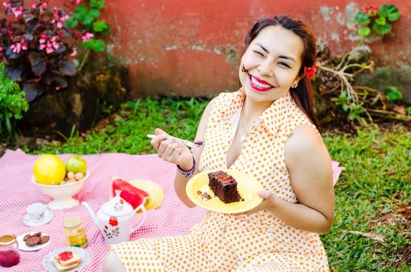 Childhood weight comments can make it hard to be an intuitive eater, but in this photo, a smiling woman with red lipstick and a retro yellow polka-dotted sleeveless dress is sitting on a pink blanket adorned with a teapot and other tea accessories, clearly enjoying a slice of chocolate cake (there's even some frosting on her face)