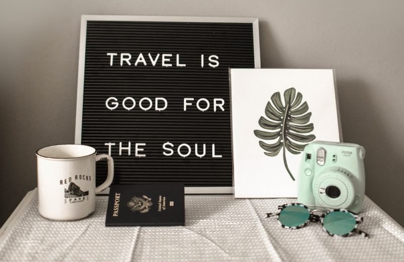 Photo of a sign that says "Travel is good for the soul" propped up on a table next to a passport, sunglasses, a mint green retro camera, a greeting card with a big tropical leaf print, and a white enameled tin coffee mug.