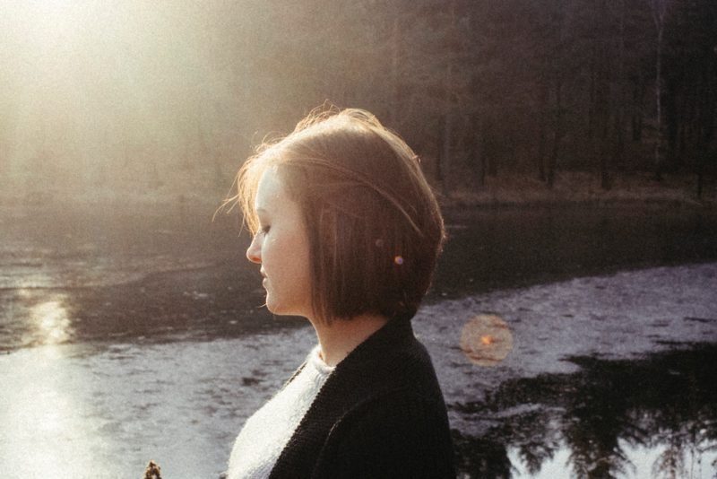 Photo of woman with a chin-length brown bob, standing near the river in diffused sunlight, looking contemplative about whether she has a problem with self-sabotage when it comes to her health.