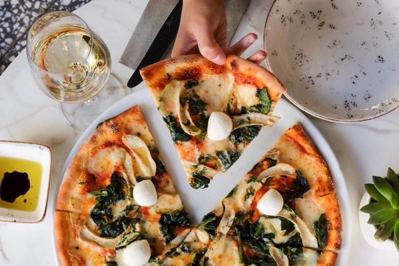 Overhead photo of a woman's hand taking a slice of thin-crust gourmet pizza. There is a glass of white wine near her hand, and a dish of olive oil and balsamic vinegar on the table, which is covered with a white tablecloth.