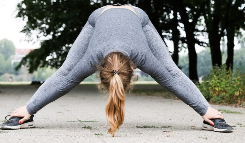 Photo of a young woman with blond hair in a ponytail, wearing heathered gray fitness leggings and a long-sleeved shirt, bent over in a wide-legged forward bend, with the back of her head towards the camera.