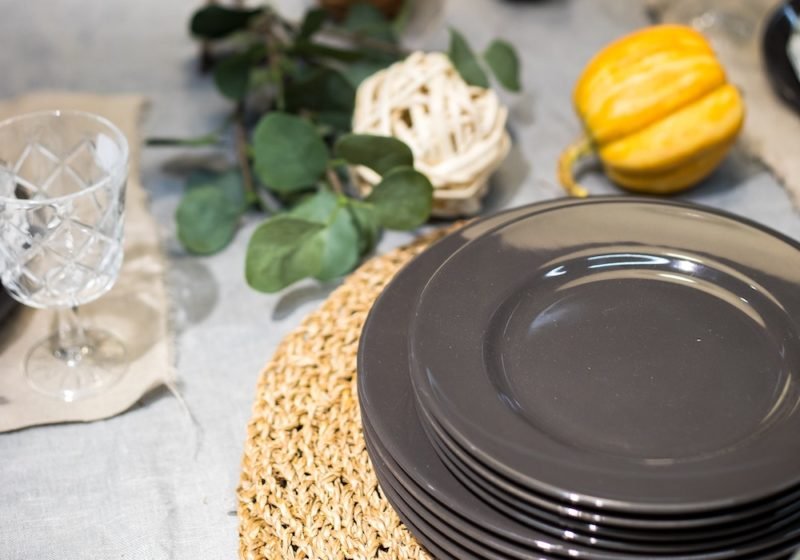Photo of holiday table, with a stack of gray plates, a cut crystal wine goblet, some greenery and a small yellow squash. Mindfulness at holiday meals can increase your happiness.