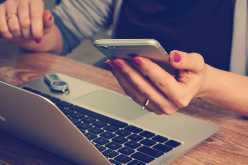 Gift yourself with more time by reducing your dependence of your smart phone and all of its alerts and notifications. Photo of a woman wearing a black top and gray cardigan sitting at a brown wooden desk with a silver-toned laptop, holding and looking at a silver smartphone.