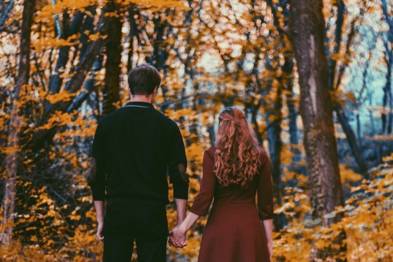 Part of honoring hunger means accepting that your food needs are different than anyone else's. Photo of a young man dressed in black and a young woman with long wavy brown hair dressed in a dark red dress, holding hands and standing with their backs to the camera, in front of woods with golden yellow foliage.