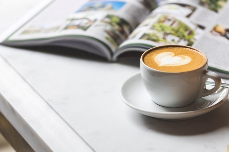 Mindful Eating Holidays: Practicing self-care, including some downtime, can make it easier to eat mindfully over the holidays. Photo of a latte with heart-shaped foam in a white cup and saucer, in front of an open magazine laying on a white tabletop.
