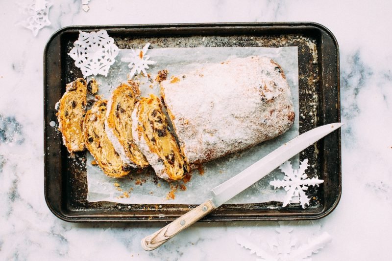 Mindful Eating Holidays: When you approach the holidays mindfully, you can enjoy holiday favorites in a balanced way. Top view photo of a partially sliced holiday fruit-and-nut loaf on parchment paper on an antique baking sheet, decorated with paper snowflakes, and sitting on a white-and-gray marble countertop.