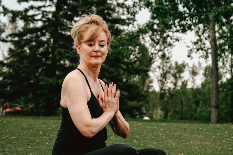 Morning rituals can include yoga and meditation, as in this photo of a middle-age woman in a black tank top and leggings, sitting cross-legged on a yoga mat in a park, with her hands together in a mediation position.