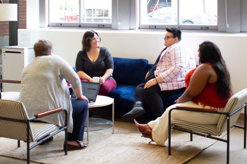 Image of four larger-bodied women sitting in chairs around a low table in an office setting.