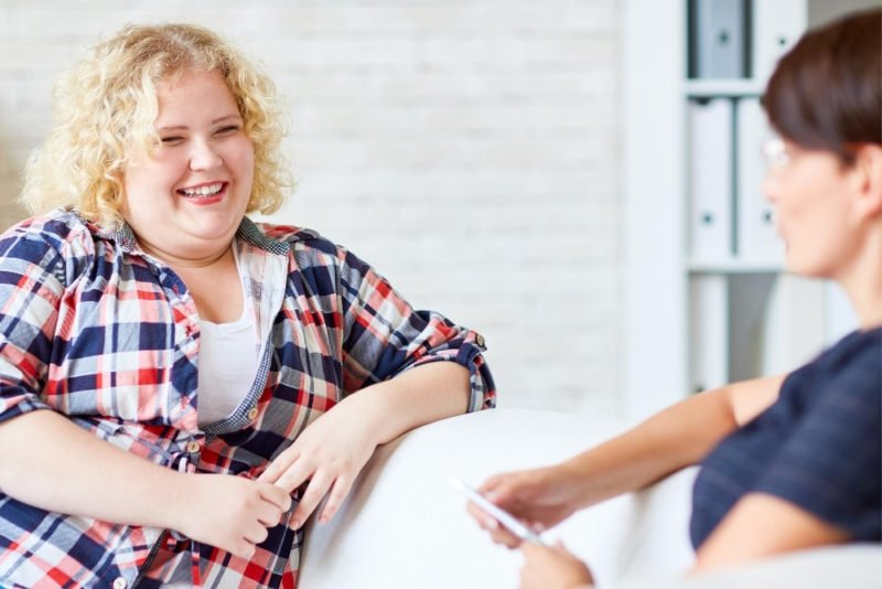A non-diet approach means making changes that support health and well being, rather than intentionally pursuing weight loss. Photos of a young woman with shoulder-length blond curly hair and a plaid shirt in a larger-body sitting on a couch and talking to a healthcare practitioner.