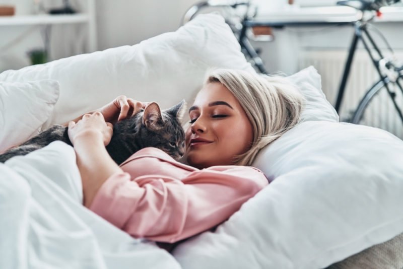The benefits of downtime include better productivity, creativity and mental health. Photo of woman in a pink pajama top snuggling with a gray tabby cat under a white duvet.