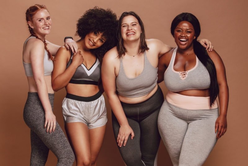 There are a lot of misconceptions about HAES, or Health at Every Size. Photo of four young women of different body sizes, smiling and wearing exercise clothing in shades of gray.