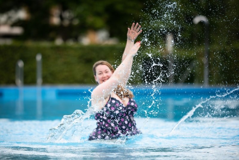 One of many HAES misconceptions is that everybody is healthy at every weight. Photo of young woman in a larger body, wearing a dark blue one-piece swimsuit with purple and pink flowers, smiling and splashing in hip-high water in a swimming pool.