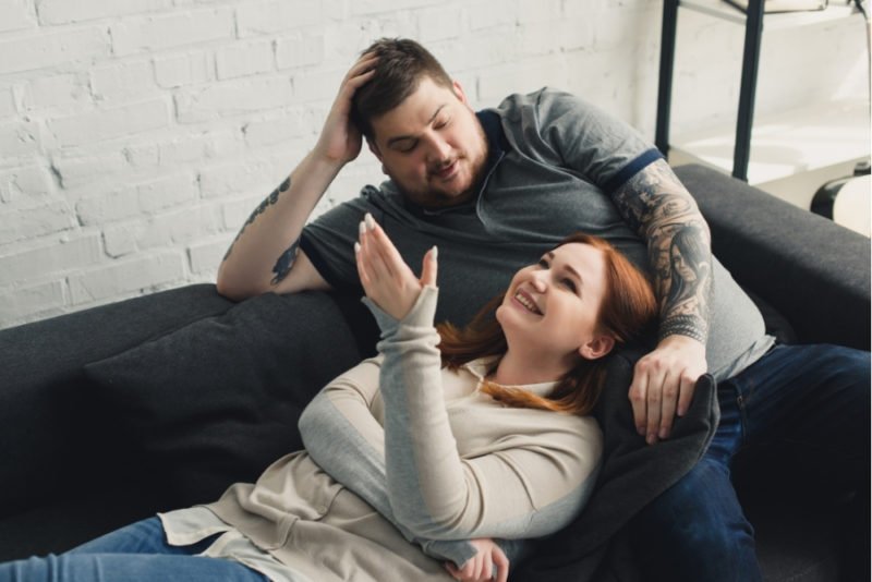 HAES points out that health is multifaceted, and includes social and emotional aspects. Photo of a young man in a larger body with arm tattoos, wearing a gray collared knit short and dark jeans, sitting reclined on a dark gray couch with a young woman with red hair, wearing a tan buttoned cardigan over a cream collared buttoned shirt and jeans, laying on the couch, smiling, with her head in his lap.