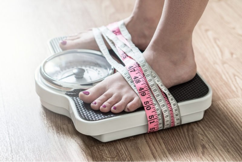 Photo is a closeup of a woman's feet standing on a white and black bathroom scale. the woman is wearing dark pink toenail polish and there are pink and white tape measures wrapped around the bottom of the scale and the top of her feet.