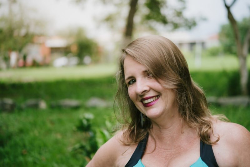 Photo of a middle-aged woman with below-shoulder length dark blond hair and dark pink lipstick, smiling and wearing a barely visible teal and black fitness tank top.