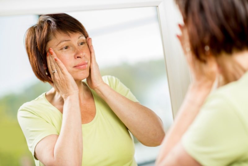 A desire to look younger may trigger an eating disorder in midlife. Photo of a middle-age woman wearing a lime green t-shirt looking at herself in a mirror with her hands on her face to smooth out wrinkles.