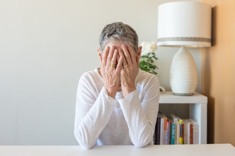 Medical complications may be worse for women who develop an eating disorder in midlife. Photo of an older women with short gray hair and wearing a long-sleeve white T-shirt sitting with her elbows on the table in front of her and her face hidden in her hands.