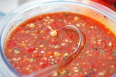 Image of a rustic gazpacho in a clear glass serving bowl, with a clear ladle, against a pale blue and white tablecloth