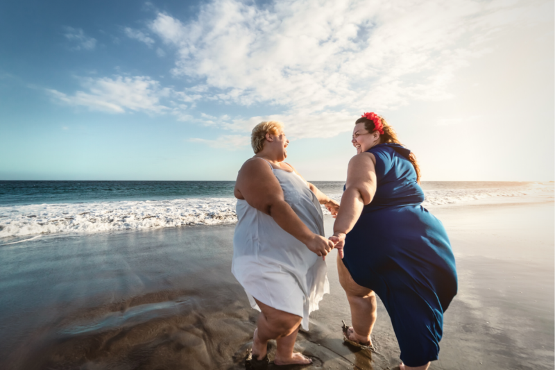 How to talk about fat bodies: Photo of two super fat women walking on the beach near the surf, holding hands and smiling. One woman has short blond hair and is wearing a white flowy sleeveless dress. The other woman has long medium brown hair and is wearing a flowy navy blue sleeveless dress and has red flowers in her hair. The sky is blue with scattered clouds.