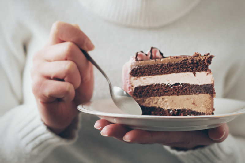Close up photo of a white woman wearing a cream-colored turtleneck sweater holding a small white plate with a slice of chocolate cake