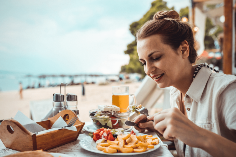 White woman eating for pleasure by enjoying Greek food at a restaurant table on the beach