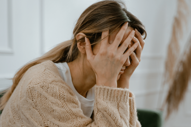 Young woman with long dark blonde hair wearing a white T shirt under a tan cable knit sweater holding her face in her hands and looking stressed or anxious.