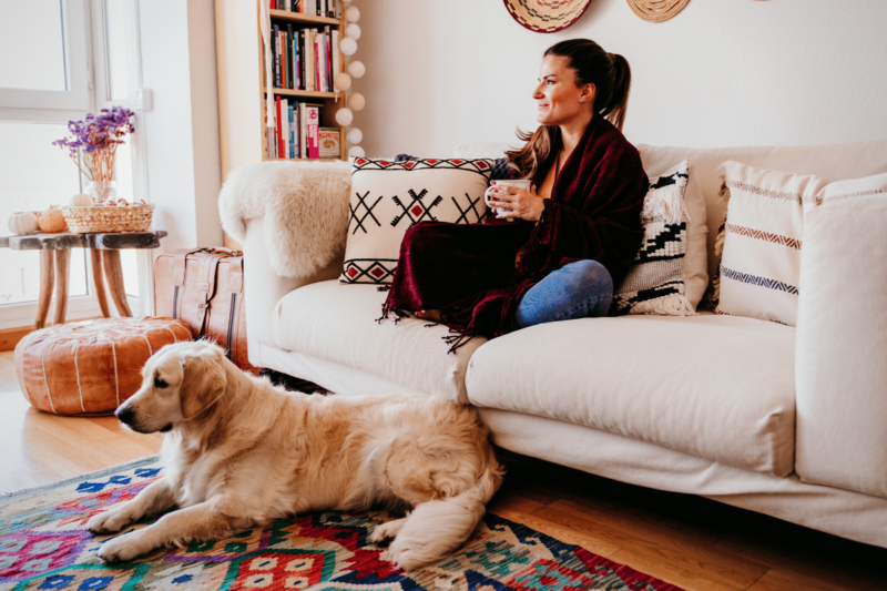 Woman with long dark hair in a ponytail sitting cross-legged on a sofa wrapped in a blanket, holding a mug of tea or coffee and smiling to herself, with a golden retriever laying at her feet.