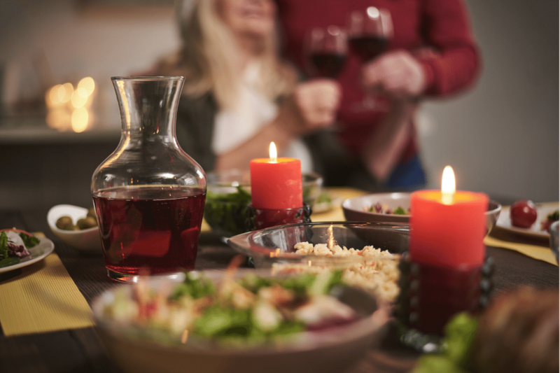 Close-up of a holiday salad with a carafe of red wine and red candles, with two out-of-focus people toasting glasses of red wine in the background