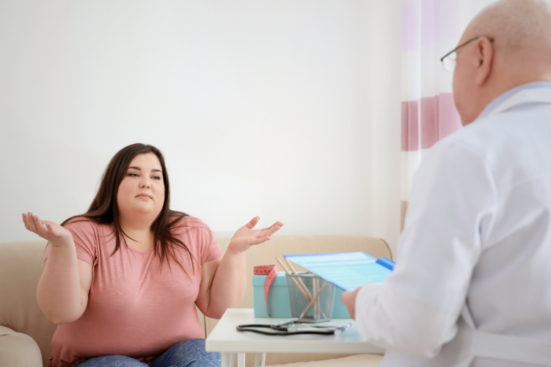 Larger-bodied woman shrugging as she sits facing a doctor in an exam room.
