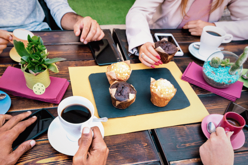 Photo of four people sitting at an outdoor table with cups of coffee. Four muffins are in the middle of the table, and only one person is reaching for a muffin.