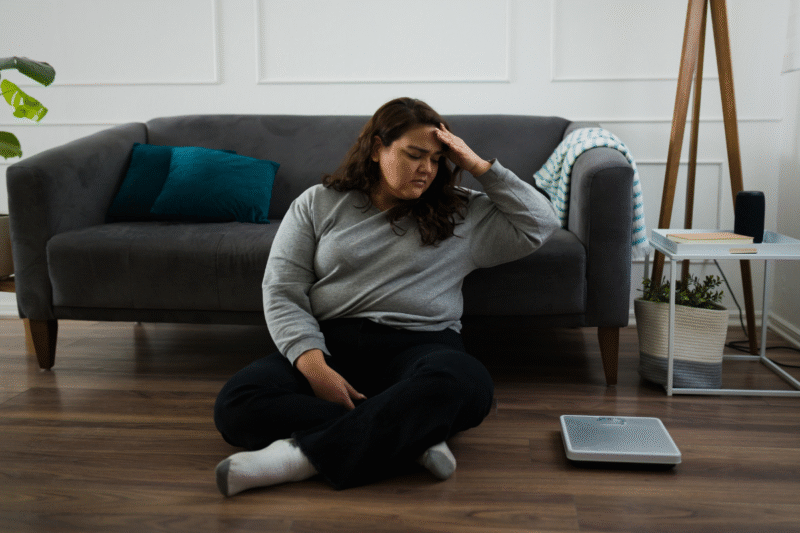Young plus-size woman sitting cross-legged on the floor next to a scale and looking upset
