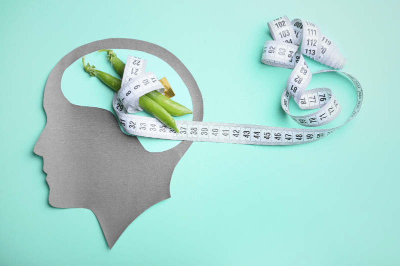 Gray paper silhouette of a face, with a tape measure wrapped around two pea pods in the space where the brain would be.