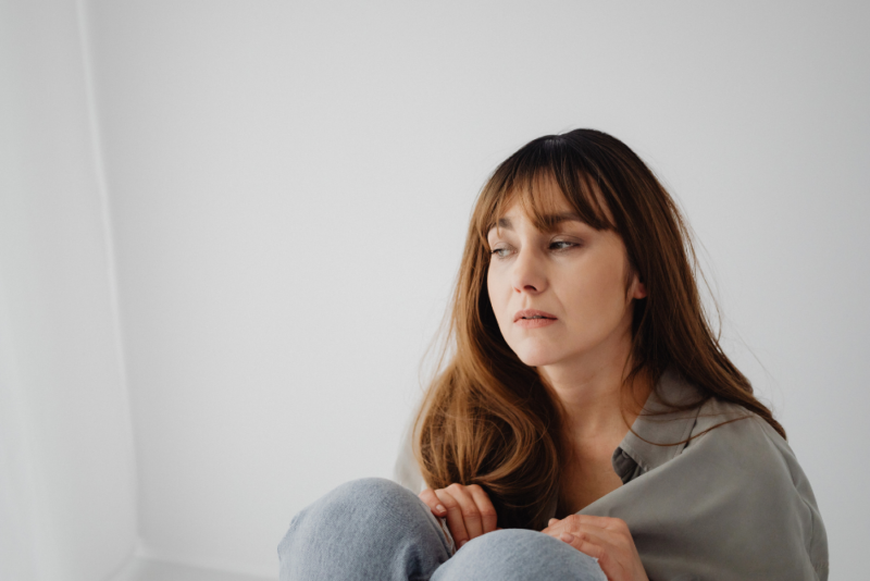 Woman with long brown hair wearing jeans and a gray button-down shirt sitting alone in her home looking sad and distressed.